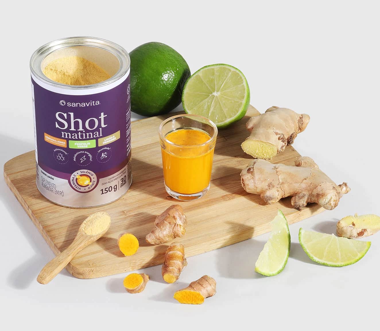 Embalagem de Shot Matinal da Sanavita ao lado de um copo com bebida laranja, fatias de gengibre e limão, e uma colher de madeira com pó amarelo, sobre uma tábua de madeira.