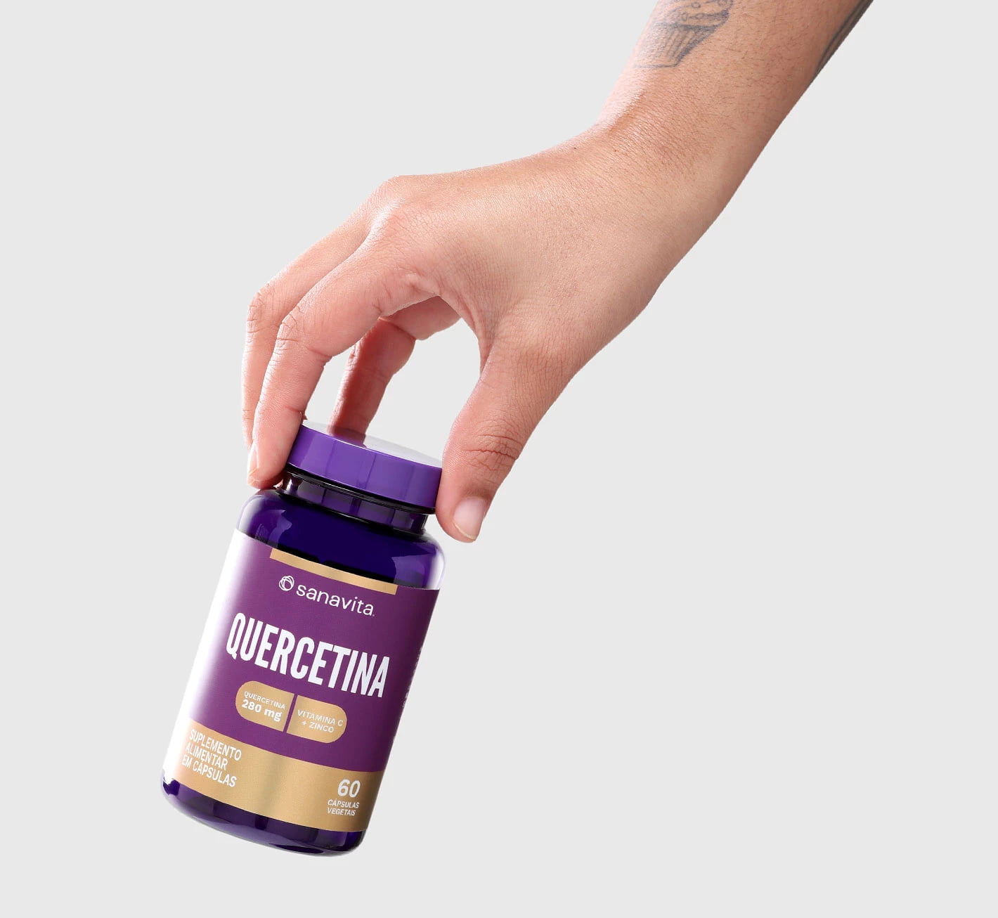 Mão segurando um frasco de suplemento alimentar Quercetina da Sanavita, com embalagem roxa e dourada, mostrando o nome do produto e as informações sobre as cápsulas.