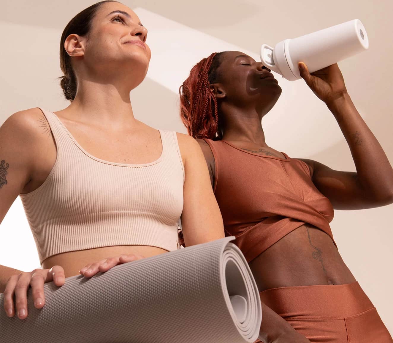 Duas mulheres em roupas de ginástica, uma segurando um tapete de yoga e a outra bebendo de uma garrafinha branca, sorrindo e com expressão de bem-estar.
