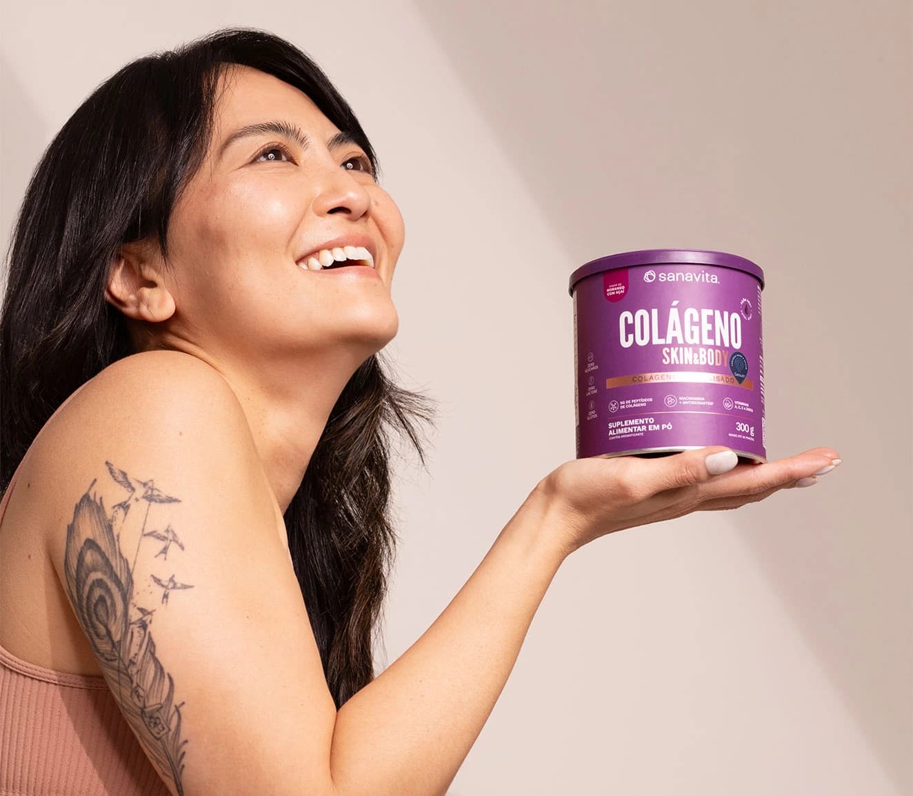 Mulher sorrindo e segurando um pote de colágeno em pó da marca Sanavita, com embalagem roxa, destacando o nome 'Colágeno Skin & Body'. Ela exibe uma tatuagem no braço esquerdo.