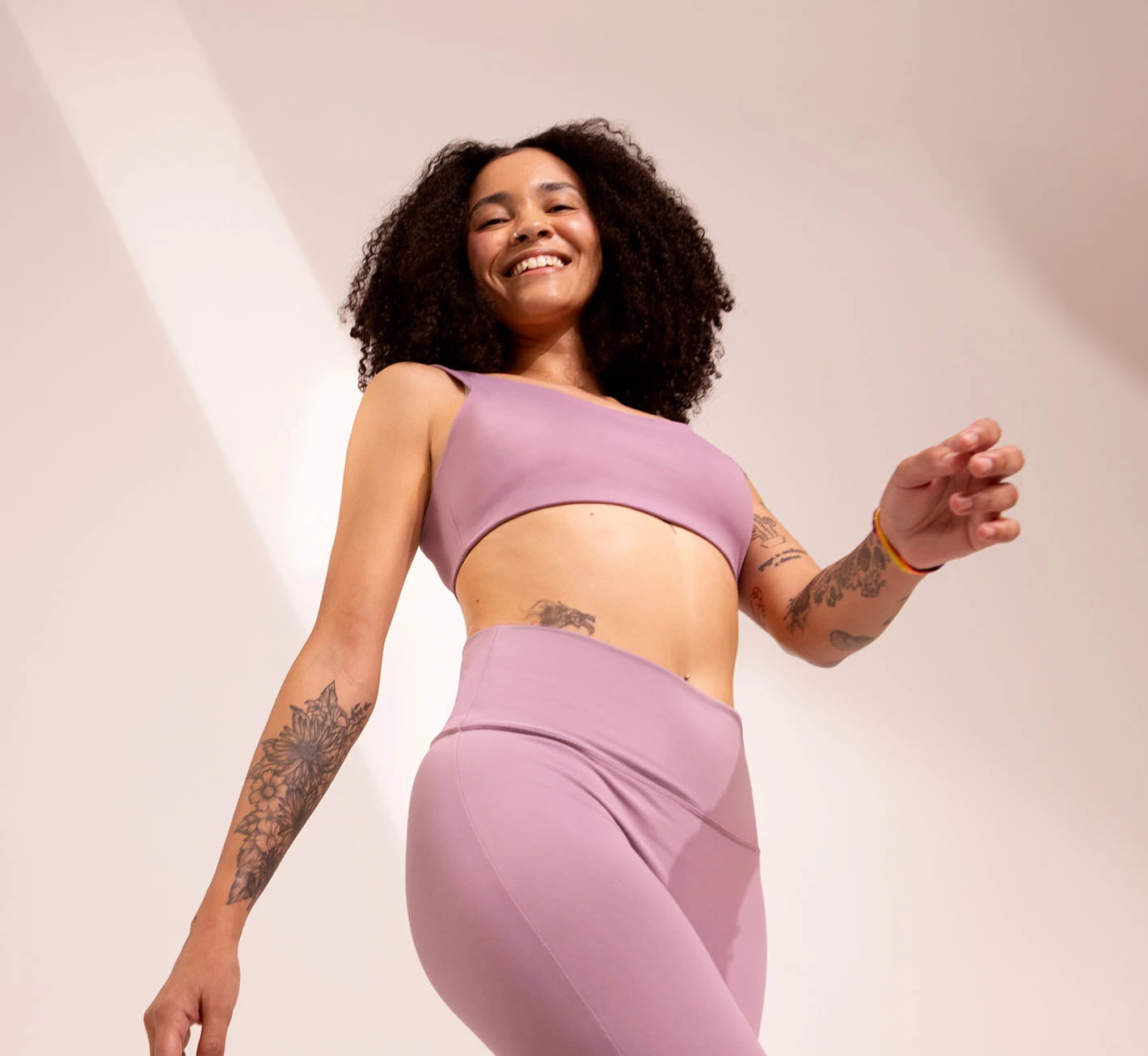 Mulher sorrindo, usando um conjunto de roupas esportivas lilás, com cabelo cacheado e tatuagens visíveis nos braços, posando de forma descontraída.